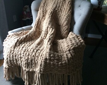 May include: A chunky knit throw blanket in a beige color with long fringe on the bottom edge. The blanket is draped over a chair.