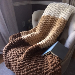 May include: A brown, beige, and white chunky knit throw blanket draped over a chair with a book on top. The blanket is made of thick yarn and has a soft, fluffy texture.