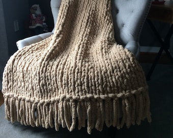 May include: A chunky knit throw blanket in a beige color with long fringe. The blanket is draped over a chair in a living room setting.