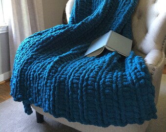 May include: A teal blue chunky knit throw blanket draped over a chair with a book on top.