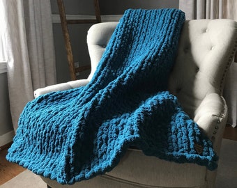 May include: A teal blue chunky knit throw blanket draped over a light gray armchair. The blanket has a textured, woven look.