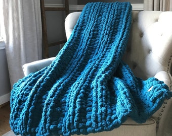 May include: A teal blue chunky knit throw blanket draped over a white armchair. The blanket has a textured, bobbly design.