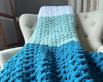 May include: A crocheted blanket with a gradient of blue, teal, and white. The blanket is made with a chunky yarn and has a textured, puffy look.