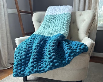 May include: A teal, light blue, and white chunky knit throw blanket draped over a beige armchair. The blanket has a textured, bobbly surface.