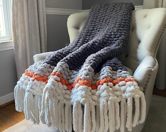 May include: A chunky knit throw blanket with a gray, white, and orange color scheme. The blanket has a fringe border and is draped over a chair.