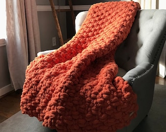 May include: A bright orange chunky knit throw blanket draped over a gray armchair. The blanket is made with a thick yarn and has a textured, puffy look.