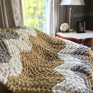 May include: A chunky knit throw blanket with a chevron pattern in shades of beige, gray, and white.
