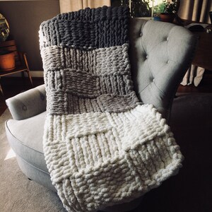 Op de afbeelding: Een dik gebreid deken in tinten grijs, beige en wit. Het deken is over een grijze fauteuil gelegd.