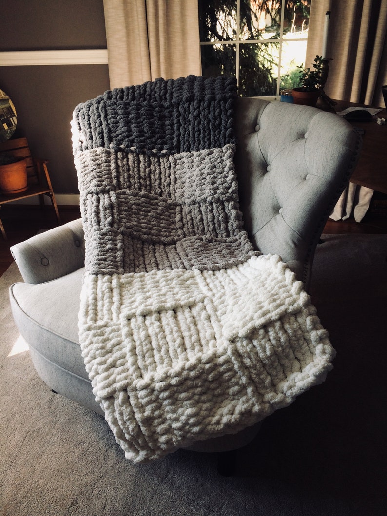 Op de afbeelding: Een dik gebreid deken in tinten grijs, beige en wit. Het deken is over een grijze fauteuil gelegd.