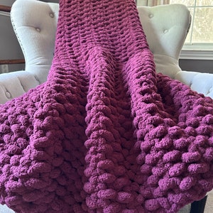 May include: A chunky knit blanket in a deep burgundy hue is draped over a white upholstered armchair. The blanket's texture is visibly plush and features a dense, textured stitch. The overall aesthetic suggests comfort and warmth, perfect for home decor.