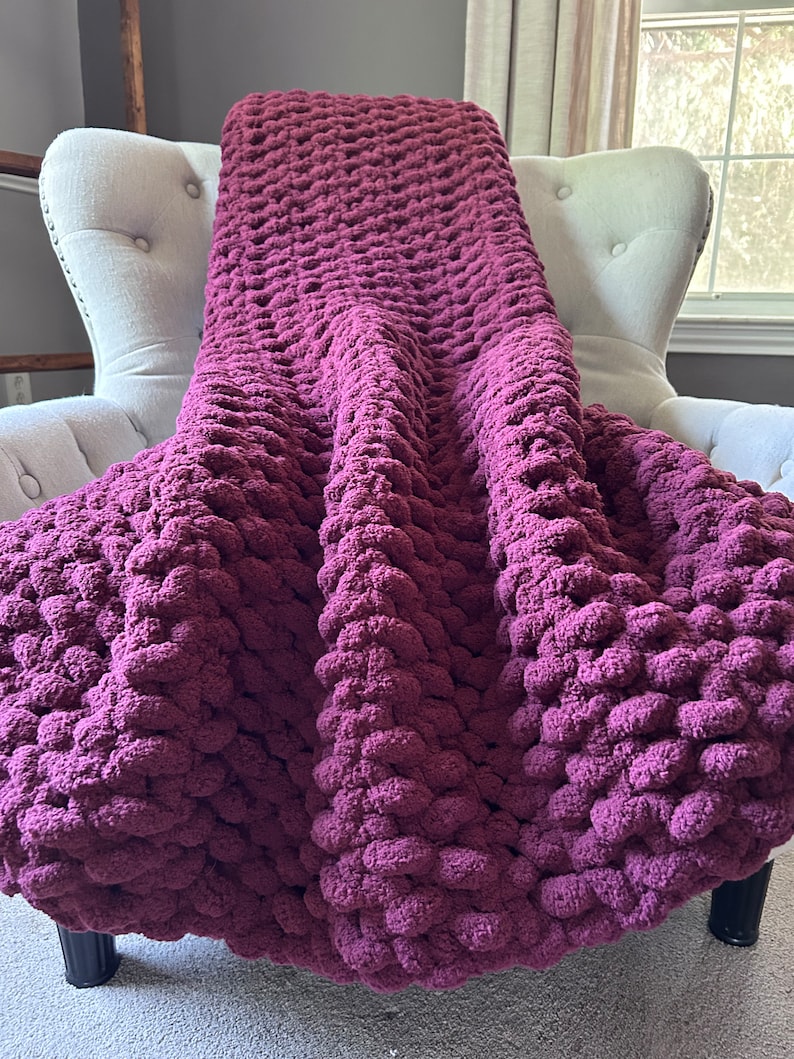 May include: A chunky knit blanket in a deep burgundy hue is draped over a white upholstered armchair. The blanket's texture is visibly plush and features a dense, textured stitch. The overall aesthetic suggests comfort and warmth, perfect for home decor.