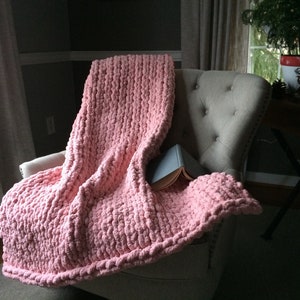 May include: A soft, pink chunky knit throw blanket draped over a chair. The blanket is made with a thick yarn and has a textured, fluffy look.