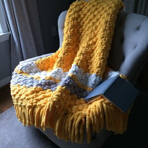 May include: A yellow and gray chunky knit throw blanket with fringe on a gray armchair. The blanket is folded over the arm of the chair and a book is lying on top of it.