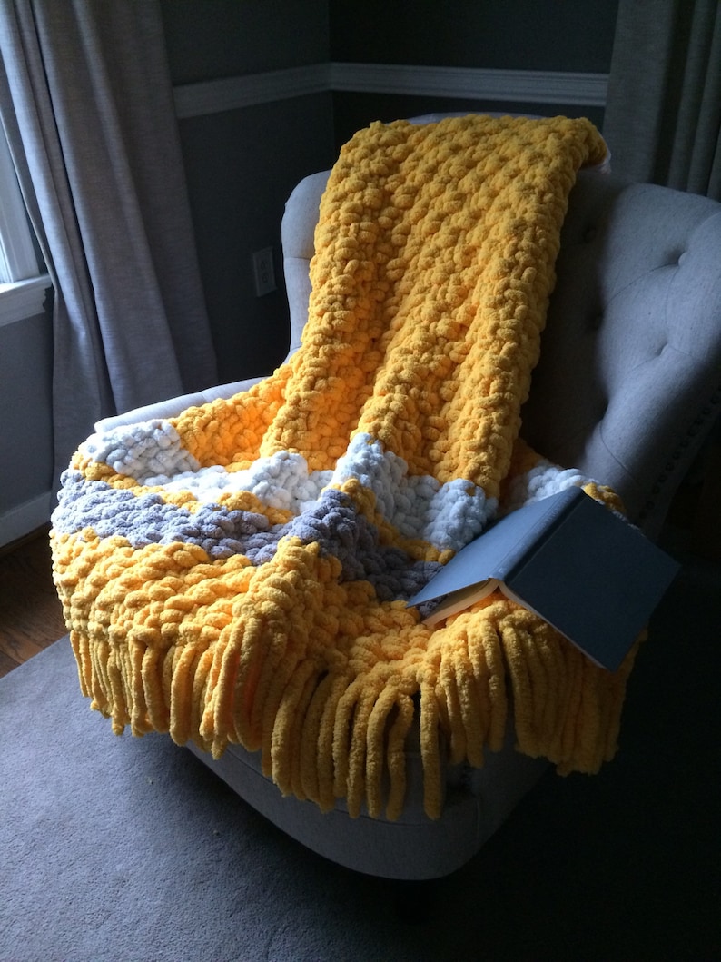 May include: A yellow and gray chunky knit throw blanket with fringe on a gray armchair. The blanket is folded over the arm of the chair and a book is lying on top of it.
