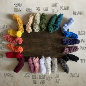 May include: A wooden board displays a color palette of yarn samples. Colors include Mustard Yellow, Khaki, Sage, Chive, Forest Green, Oceanside, Periwinkle, Classic Blue, Navy Blue, Smoky Amethyst, Onyx, Charcoal, and more. Each color is labeled.