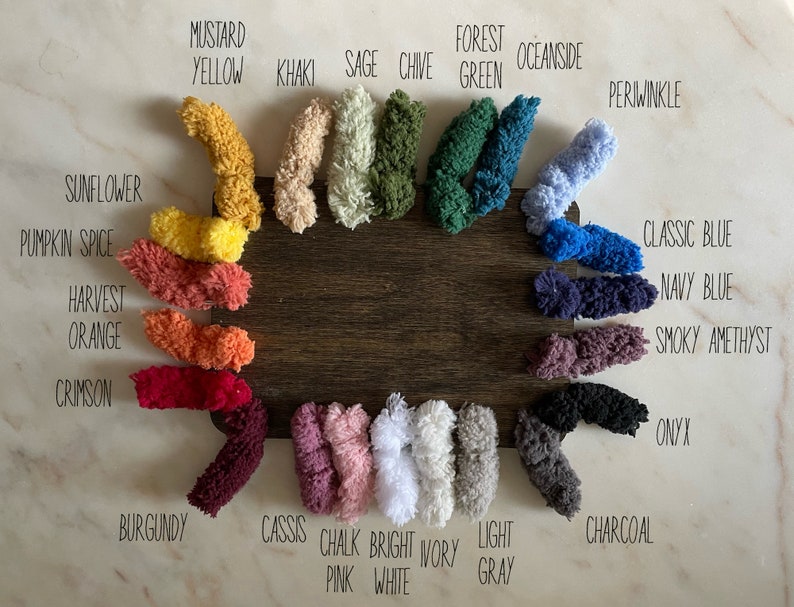 May include: A wooden board displays a color palette of yarn samples. Colors include Mustard Yellow, Khaki, Sage, Chive, Forest Green, Oceanside, Periwinkle, Classic Blue, Navy Blue, Smoky Amethyst, Onyx, Charcoal, and more. Each color is labeled.