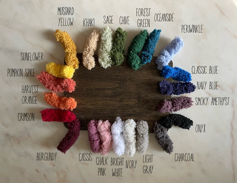 Op de afbeelding: Een kleurenkaart met garenstalen met namen zoals "Mosterdgeel", "Salie", "Bosgroen", "Zeezijde", "Blauwe druif", "Klassiek blauw", "Donkerblauw", "Rokerige amethist", "Onyx", "Antraciet", "Cassis", "Krijt roze", "Helder wit", "Lichtgrijs", "Bordeaux", "Krimzon", "Oogst oranje", "Pompoenspecerij", en "Zonnebloem".
