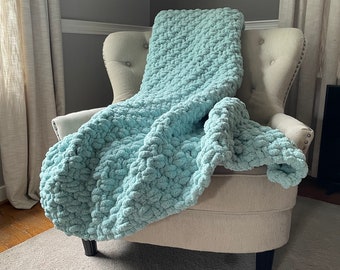 May include: A light blue chunky knit throw blanket draped over a beige armchair. The blanket has a soft, fluffy texture.