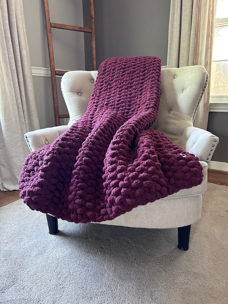 May include: A plush, burgundy-colored blanket draped over a cream-colored armchair. The blanket has a chunky knit texture, creating a cozy and inviting appearance. The chair is set against a neutral-toned wall, with a wooden ladder and curtains in the background.