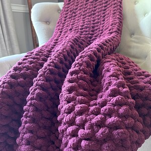 May include: A plush, burgundy-colored blanket draped over a white armchair. The blanket has a chunky, textured knit design, creating a cozy and inviting appearance. The deep purple hue adds warmth and visual interest to the scene, suggesting comfort and relaxation.