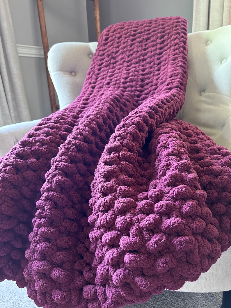May include: A plush, burgundy-colored blanket draped over a white armchair. The blanket has a chunky, textured knit design, creating a cozy and inviting appearance. The deep purple hue adds warmth and visual interest to the scene, suggesting comfort and relaxation.