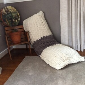 May include: A large, white and gray knitted pillow with a chunky knit texture. The pillow is shaped like a beanbag chair and is sitting on a gray rug in a room with gray walls.