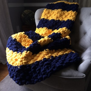 May include: A striped crocheted throw blanket in navy blue and yellow. The blanket is made with chunky yarn and has a fluffy texture. It is draped over a gray armchair.