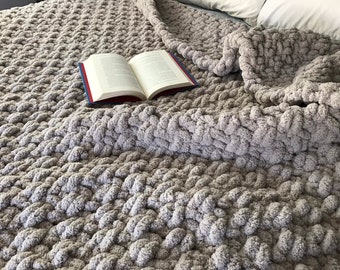 May include: A soft, gray, chunky knit throw blanket with a book resting on top. The blanket is made with a thick yarn and has a textured, woven pattern.
