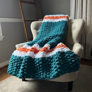May include: A teal, white, and orange chunky knit throw blanket draped over a beige armchair. The blanket has a textured, bobbly surface.