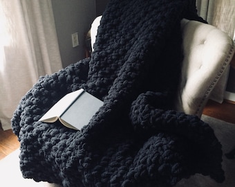 May include: A black chunky knit throw blanket draped over a white armchair with a book resting on top.