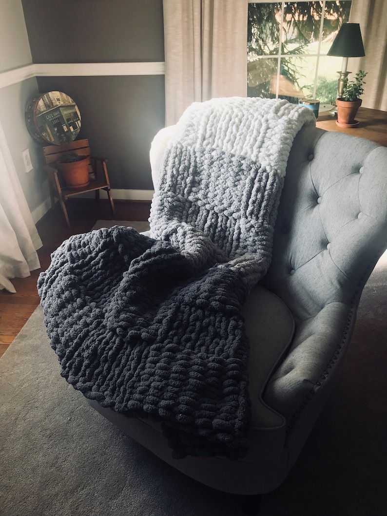 Op de afbeelding: Een grijze fauteuil met een grof gebreide deken in grijstinten en wit. De deken is over de rugleuning van de stoel gelegd en hangt aan de zijkanten.