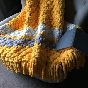 May include: A yellow, gray, and white striped chunky knit throw blanket with fringe on a gray armchair. The blanket is folded over the arm of the chair and a book is lying on top of it.