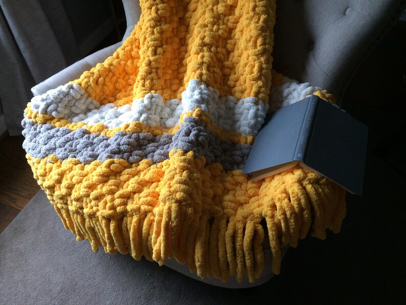 May include: A yellow, gray, and white striped chunky knit throw blanket with fringe on a gray armchair. The blanket is folded over the arm of the chair and a book is lying on top of it.