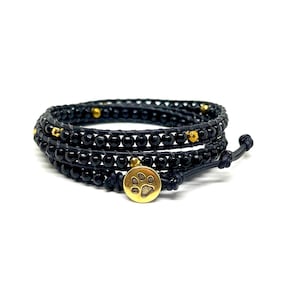 May include: A black beaded wrap bracelet with a gold paw print charm. The bracelet is made of three strands of black beads and has a gold clasp.