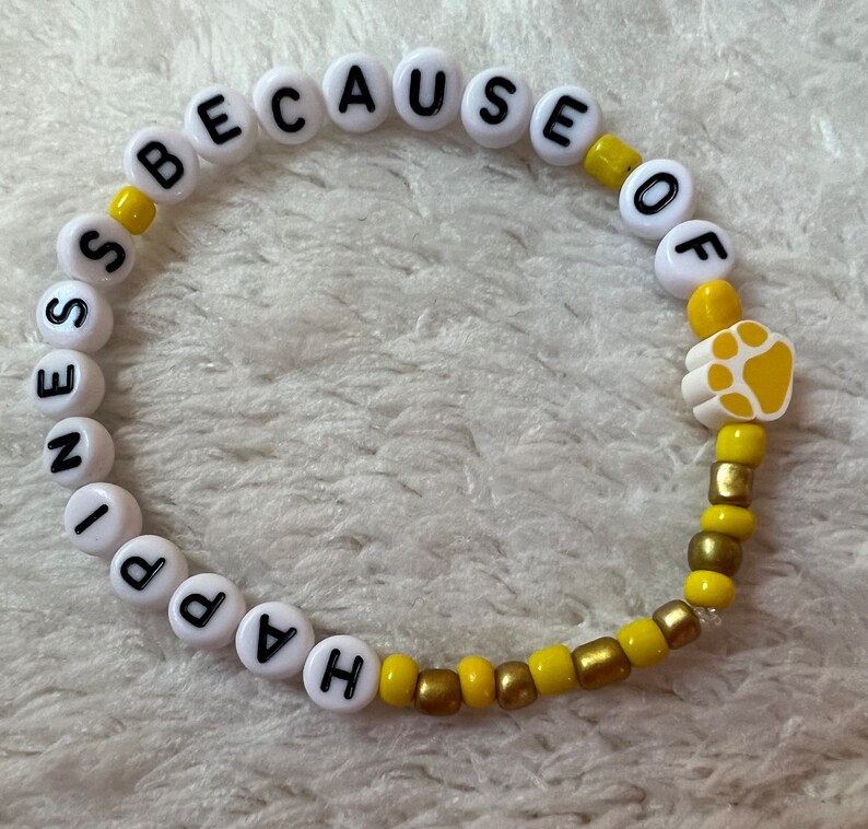 Puede incluir: Un brazalete de cuentas blancas y amarillas con las palabras "Happiness because of" y un dije de huella de pata.