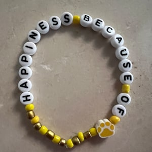 Puede incluir: Pulsera de cuentas blancas con las palabras "Happiness because of" y un dije de pata amarilla. Las cuentas alternan entre blanco y amarillo.
