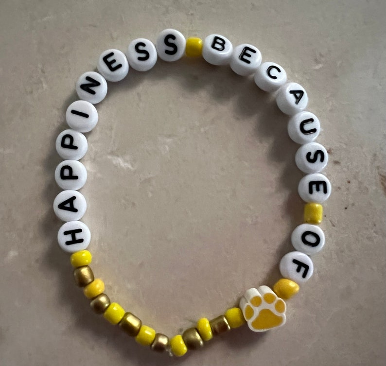 Puede incluir: Pulsera de cuentas blancas con las palabras "Happiness because of" y un dije de pata amarilla. Las cuentas alternan entre blanco y amarillo.