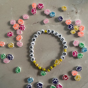 Puede incluir: Un brazalete blanco de cuentas con las palabras "Happiness because of" escritas en letras negras. El brazalete est&aacute; rodeado de abalorios con forma de huella de pata de colores.