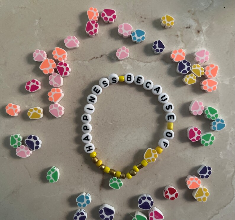 Puede incluir: Un brazalete blanco de cuentas con las palabras "Happiness because of" escritas en letras negras. El brazalete est&aacute; rodeado de abalorios con forma de huella de pata de colores.