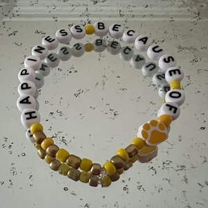 Puede incluir: Un brazalete de cuentas blancas y amarillas con las palabras "Happiness because of" y un dije con forma de huella de pata.