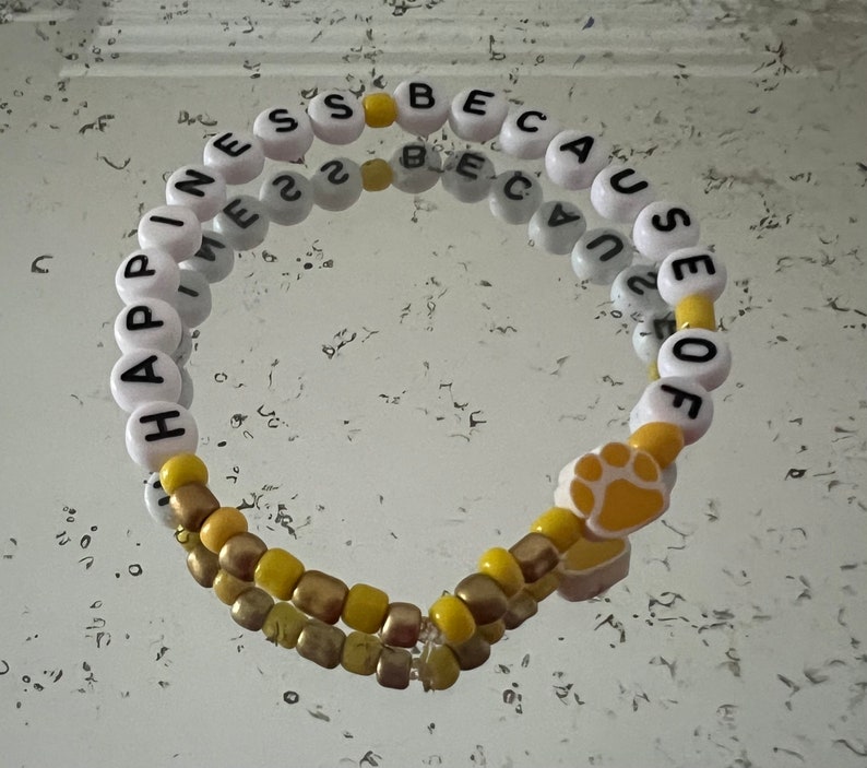 Puede incluir: Un brazalete de cuentas blancas y amarillas con las palabras "Happiness because of" y un dije con forma de huella de pata.