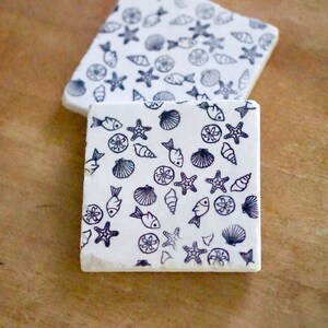 May include: Two square white coasters with a repeating pattern of black sea creatures, including fish, starfish, and seashells. The coasters are made of a light-colored material and rest on a wooden surface.