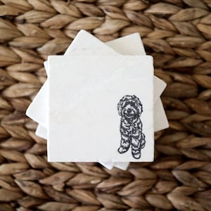 May include: A stack of square, white marble coasters. Each coaster features a black and white illustration of a dog. The coasters are stacked on a woven, brown surface.