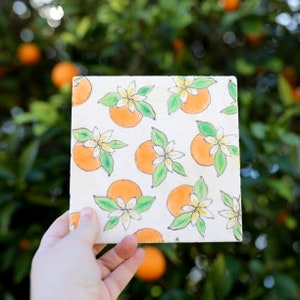 May include: A square ceramic tile featuring a repeating hand-painted pattern of bright orange citrus fruits, white blossoms, and green leaves on an off-white background. The design is rendered in a watercolor style with visible ink outlines.