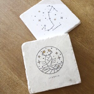 May include: Two white square coasters with black and gold Scorpio zodiac signs. One coaster features a constellation of stars and the word "SCORPIUS". The other coaster features a stylized scorpion with the word "SCORPIO".