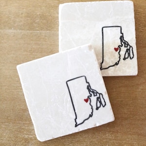 May include: Two white square coasters with a black outline of the state of Rhode Island and a red heart in the center.