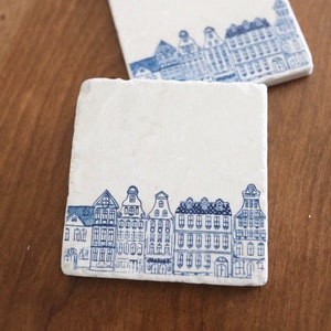 May include: Two white ceramic coasters with a blue and white illustration of a row of houses. The coasters are on a wooden surface.