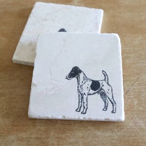 May include: Two square, off-white marble coasters. Each coaster features a black and white illustration of a dog. The dog has a black head and a black spot on its side. The coasters are on a wooden surface.