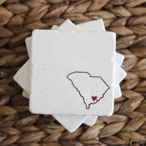 May include: A set of four white marble coasters with a black outline of the state of South Carolina and a red heart in the lower right corner of the state.