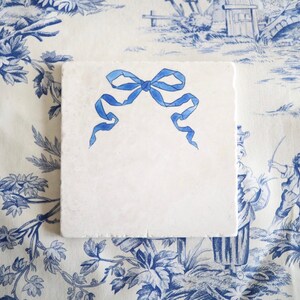 May include: A square, off-white stone coaster featuring a hand-painted blue ribbon bow centered on its surface. The coaster has a smooth, flat top and slightly textured, natural stone edges, providing a clean and simple aesthetic for the home.